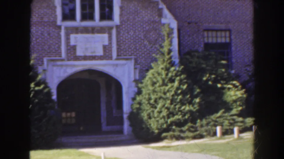 1941 Florida home-movie frame showing a formal brick building entry with white trim, evergreen shrubbery, lawn, and walkway.