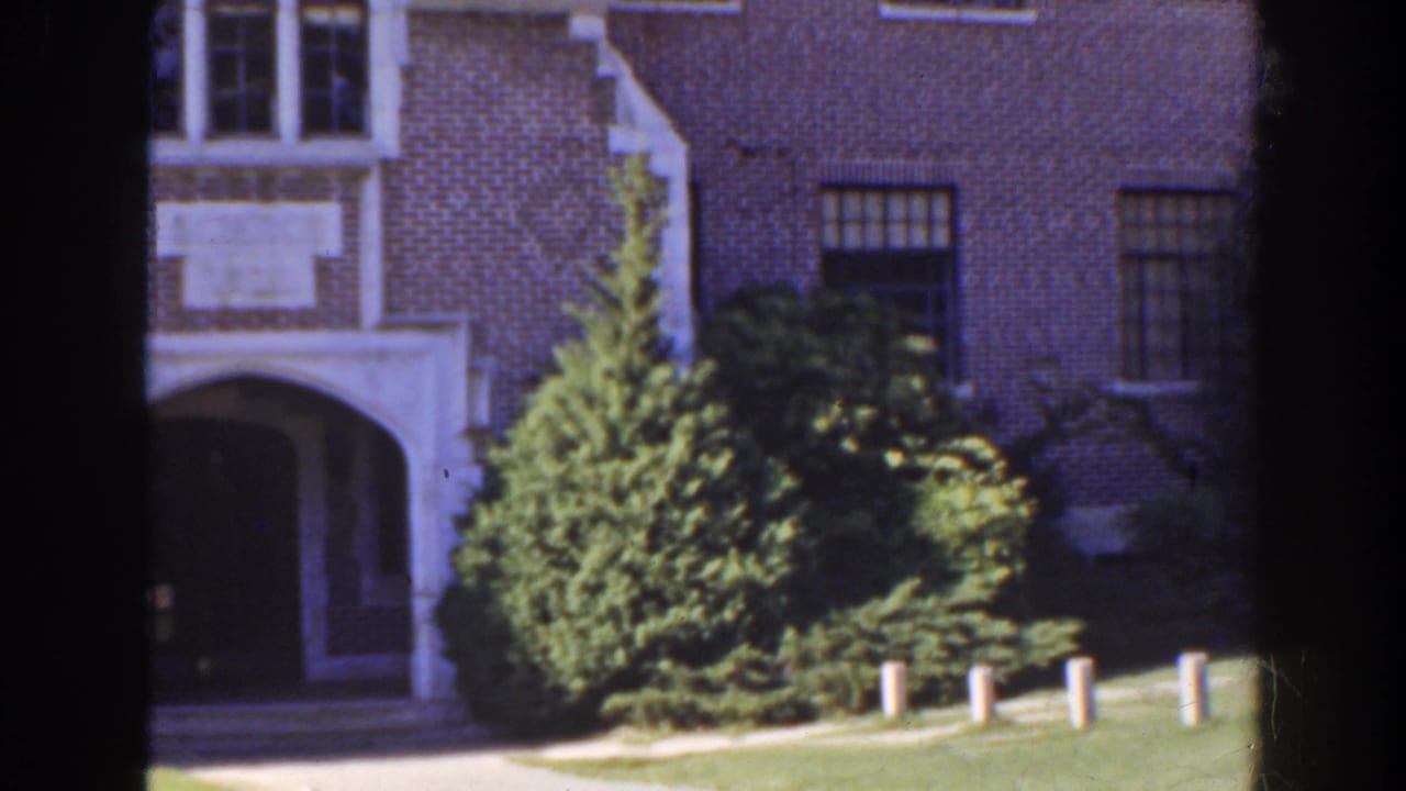 Brick house facade with arched entry, windows, shrubs, lawn, and white posts in sunlight.