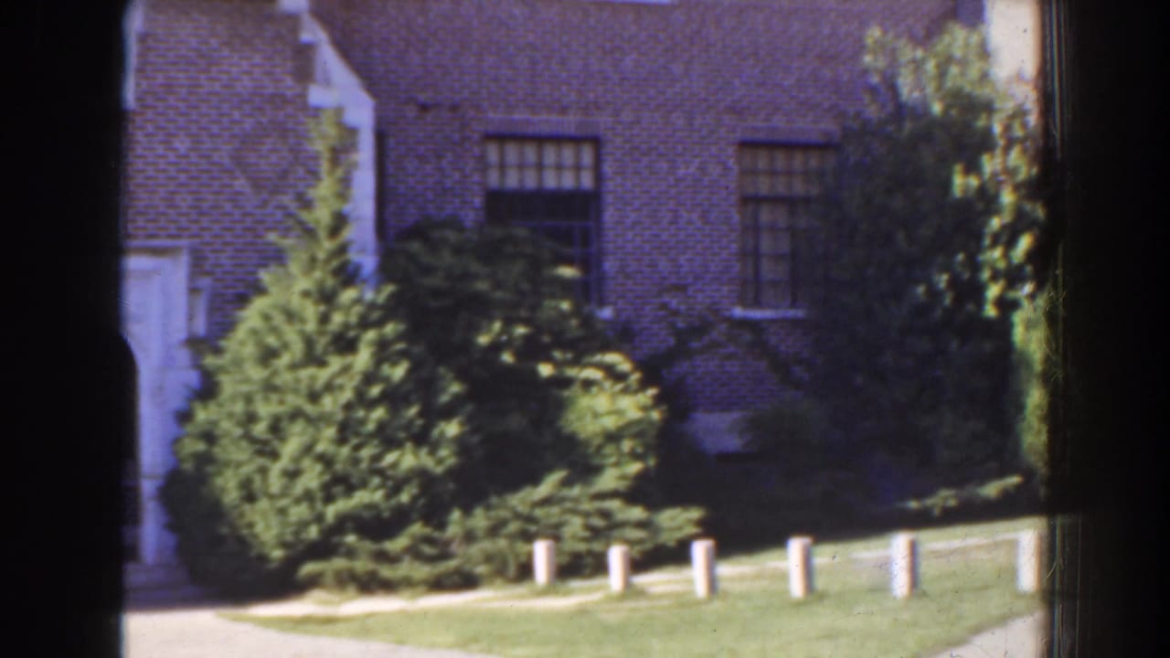 Brick house wall, barred or multi-paned windows, shrubs, grass, and white posts during a pan.