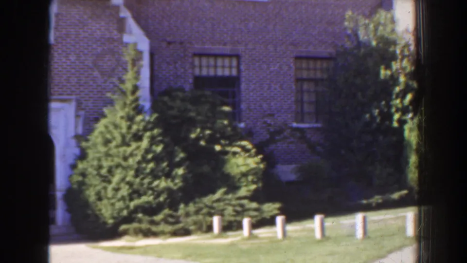 1941 Florida home-movie frame with brick house windows, sunlit shrubs, trimmed grass, and small white posts along the foreground.