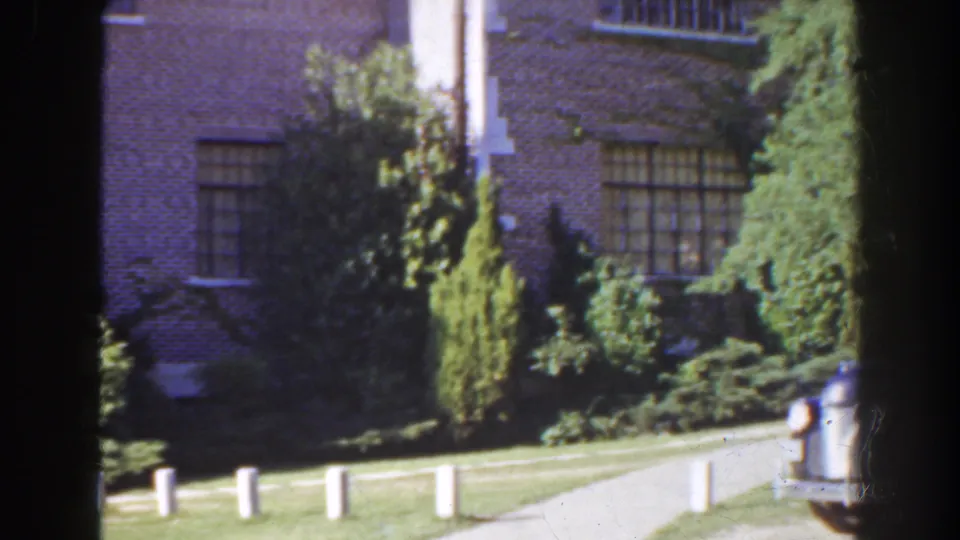 1941 Florida home-movie frame with brick windows, dense shrubs, lawn, white posts, paved path, and a partial vehicle edge at right.