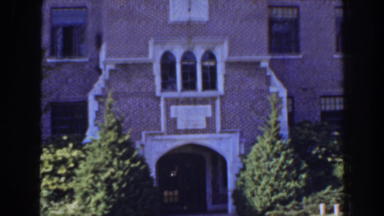 Brick building entrance with white stone trim, arched windows, trees, and shrubs.