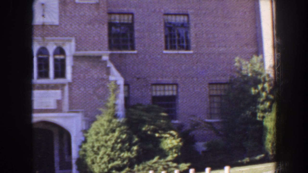 Brick building facade with arched entry, windows, shrubs, and a narrow lawn area.