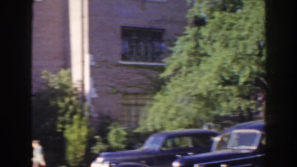 1941 Florida home-movie frame with brick building windows, dense greenery, and two dark rounded automobiles in the foreground.