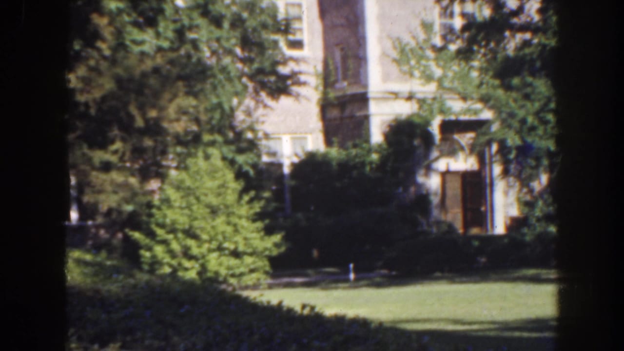 Large pale building partly obscured by trees and shrubs across a green lawn.