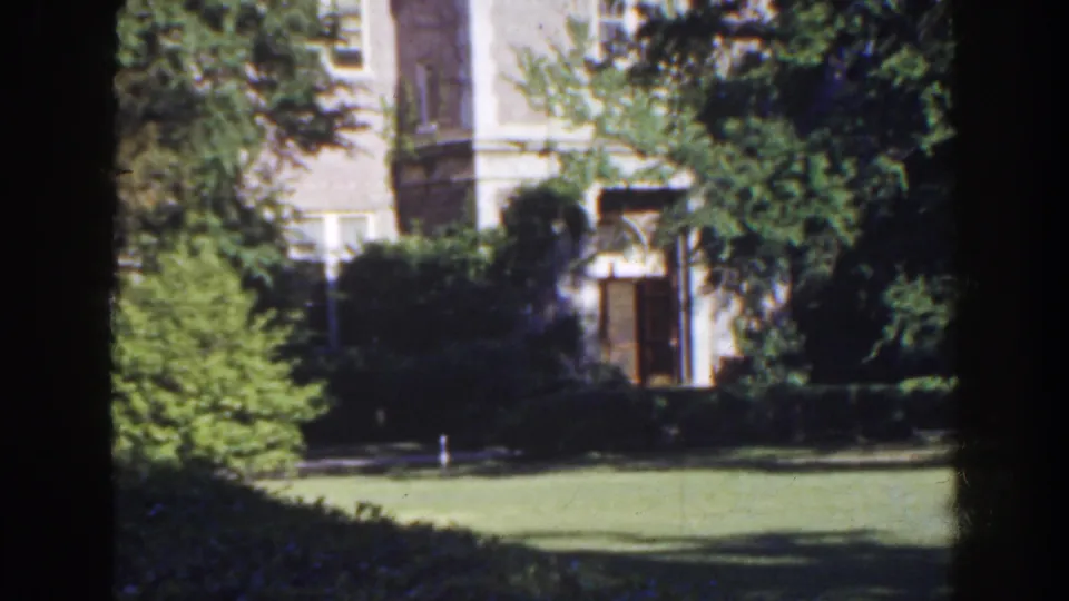 Vintage 8mm frame of a Florida property exterior, with a pale facade filtered through trees and shrubbery.