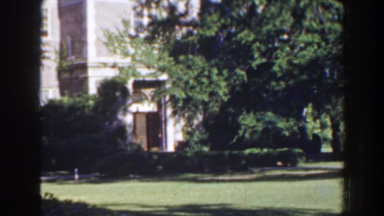 White stone building partly hidden behind trees, lawn, hedges, and dark film-frame borders.