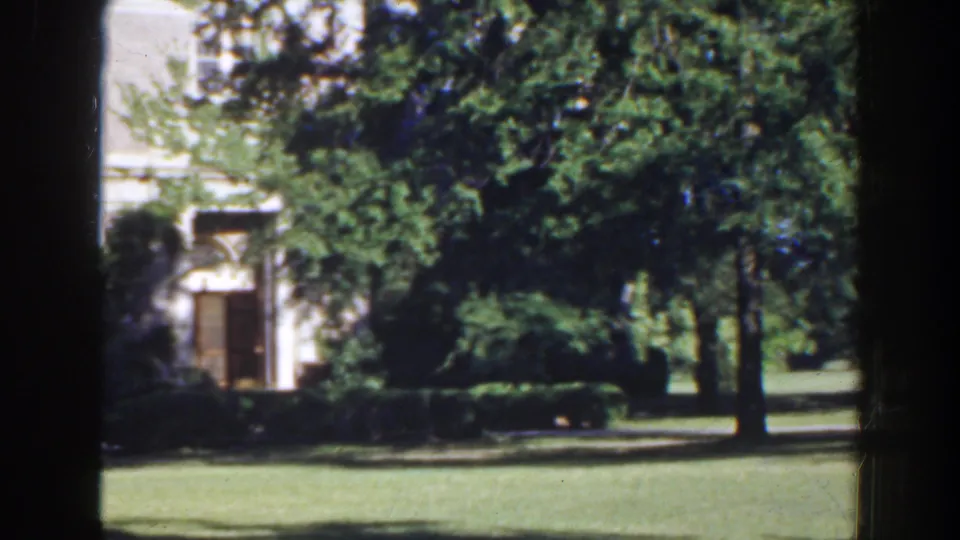 1941 Florida 8mm frame with a pale building and doorway partly visible behind dense trees, trimmed hedges, and open grass.