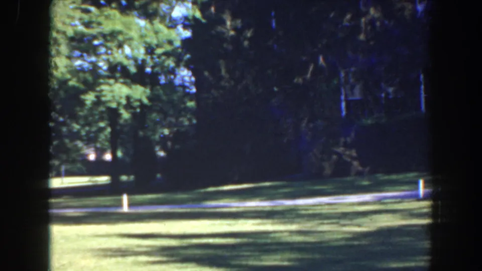 1941 Florida home-movie frame with a shaded tree line, bright lawn, pale path, and a small white post near the route.