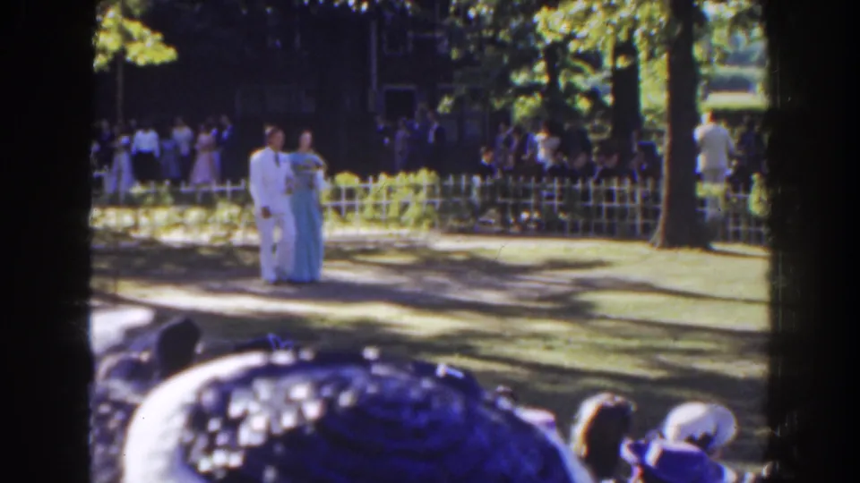 In 1941 Florida home-movie footage, a man in a white suit and a woman in a light blue gown walk past a fence and watching crowd.
