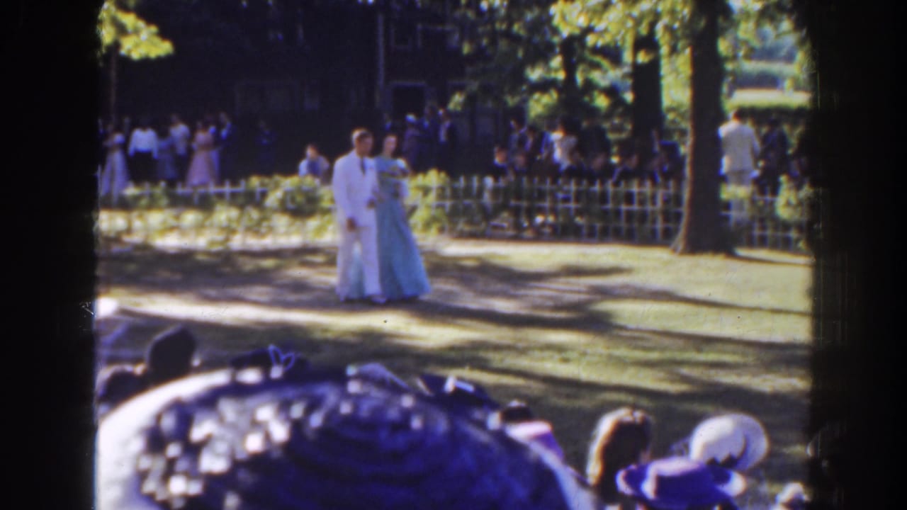 Formal couple continues along the garden aisle with crowd behind a fence.