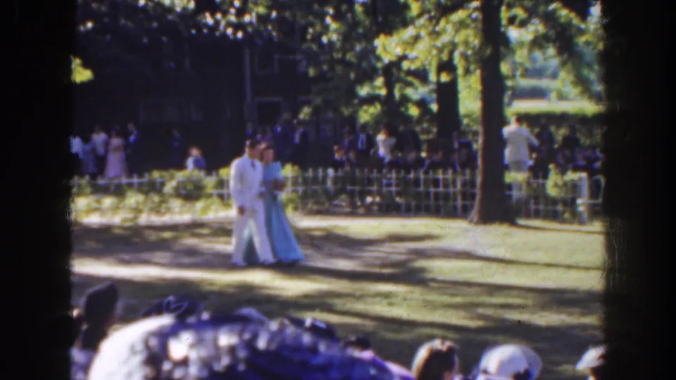 A man in a white suit and woman in a pale blue gown continue through the Florida garden scene as spectators stand and sit nearby.