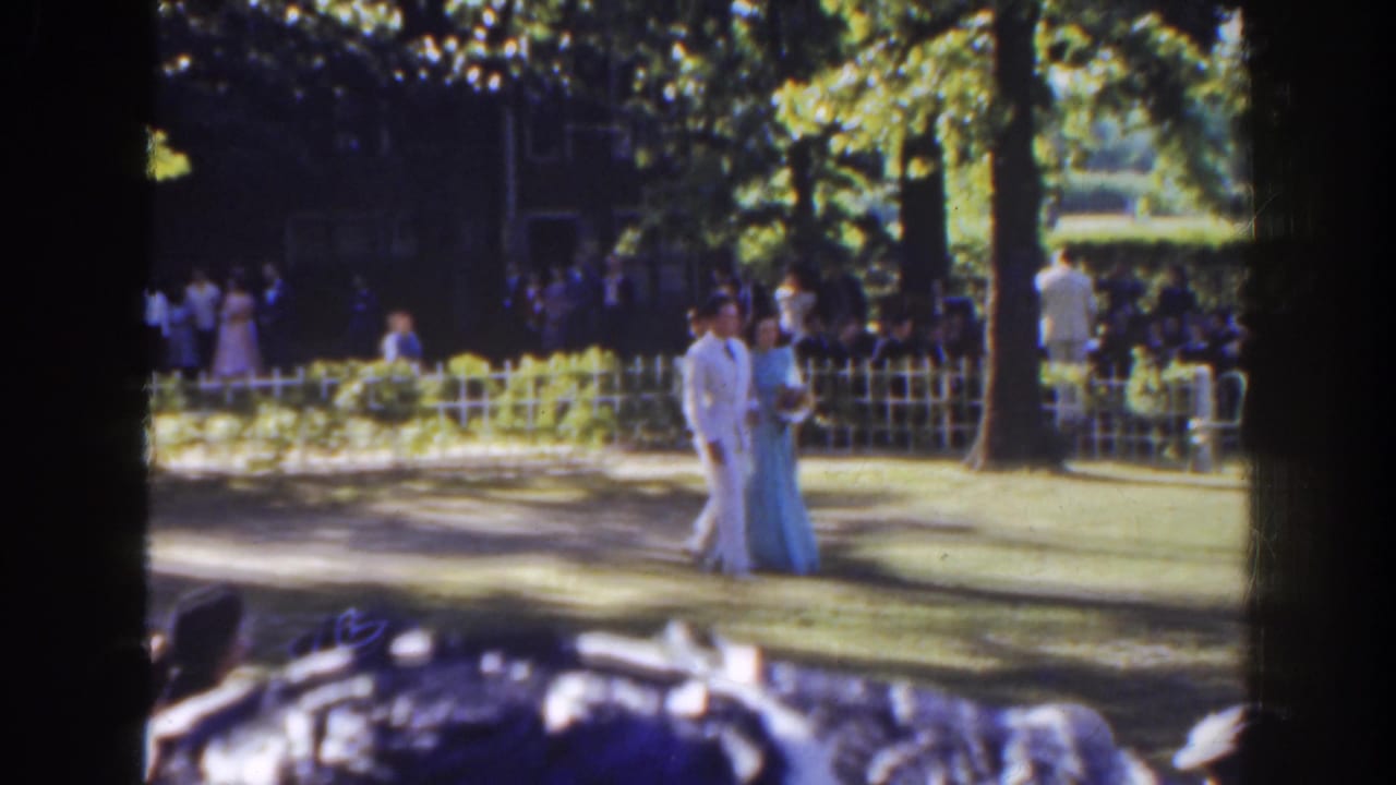 Formal couple walking across a shaded lawn with guests behind a white fence.