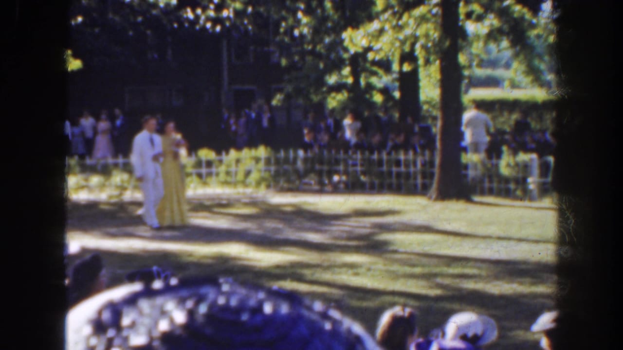 Couple in formal clothing walking near trees and guests in a park setting.