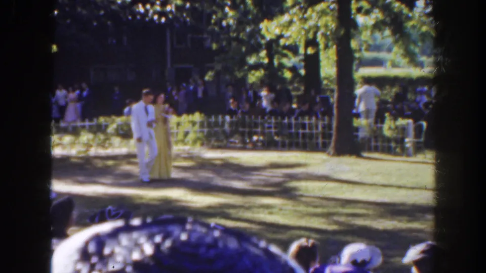 A man in a white suit and a woman in a long light gown move through the park ceremony area, with guests and hats visible beyond the fence.