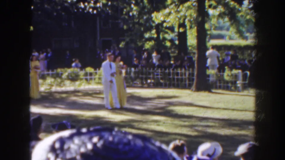 Near the end of the clip, the couple pauses or slows on the shaded lawn while the outdoor wedding guests remain visible behind the fence.