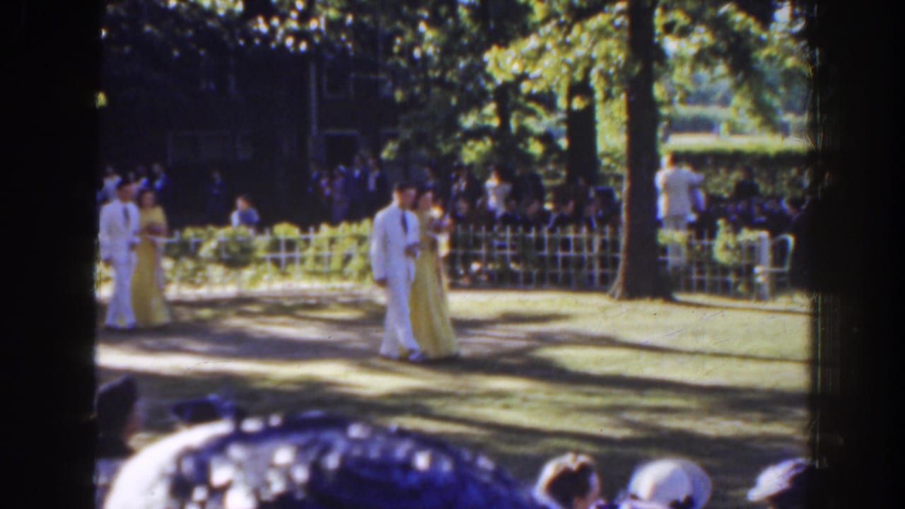 Couple in formal dress walks across a shaded lawn before seated spectators.