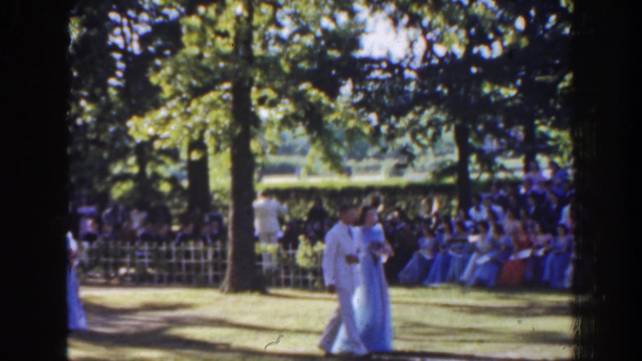 Couple in light formal clothing passes in front of a seated outdoor audience.