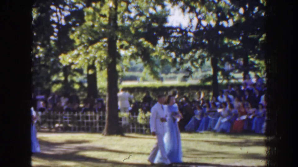 A formally dressed couple walks through a sun-dappled garden setting in Florida, with rows of spectators visible behind them.