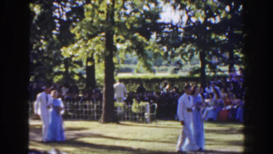 Two couples in white suits and pastel gowns appear in the garden procession, with spectators seated along the fence line.