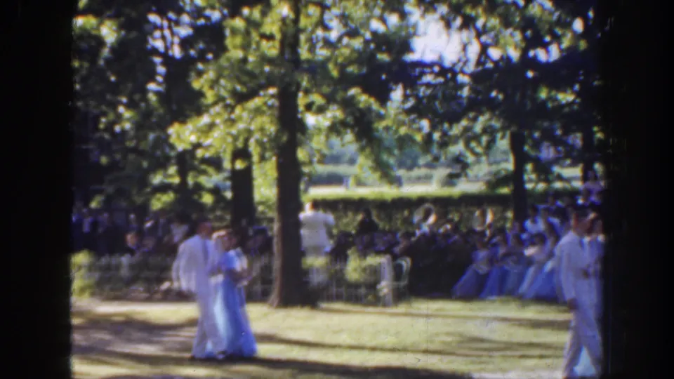 A couple in light formal clothing appears near center frame, with seated spectators and another white-suited man at the edge.