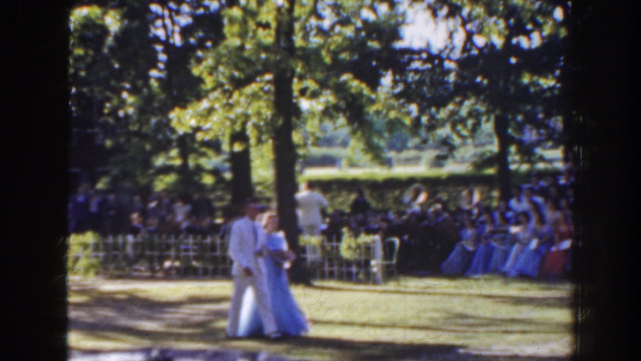 Couple in light formal clothing walks across a lawn as guests sit behind a picket fence.