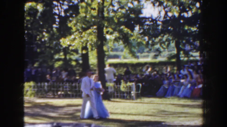 In 1941 Florida footage, a formally dressed couple crosses a sunlit garden lawn with seated guests behind a white fence.