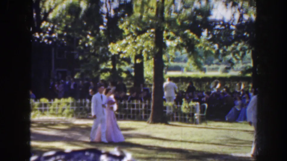 1941 Florida home-movie frame showing a formally dressed couple walking through an outdoor wedding setting with guests behind a low fence.