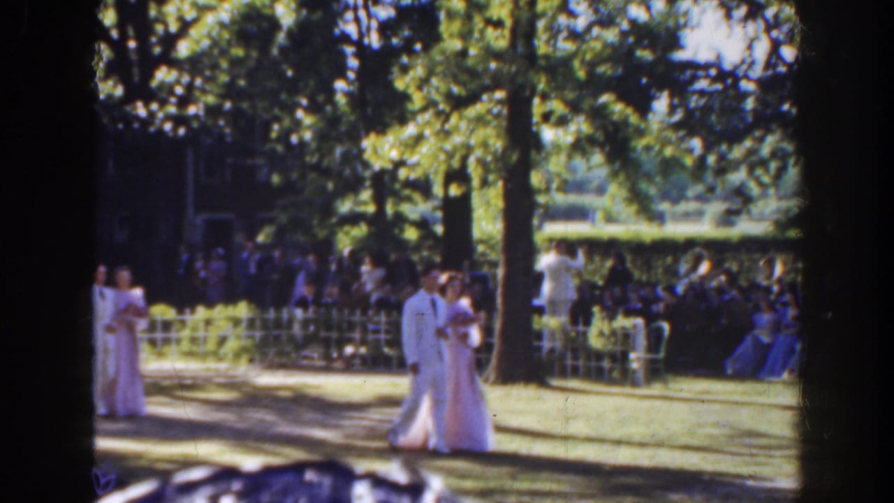 Formal wedding couple walking past guests seated under trees in a garden.