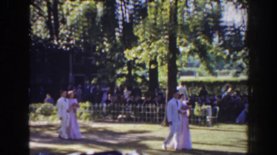 A 1941 Florida wedding procession continues as the couple crosses the lawn and a bridesmaid follows near the guest seating.