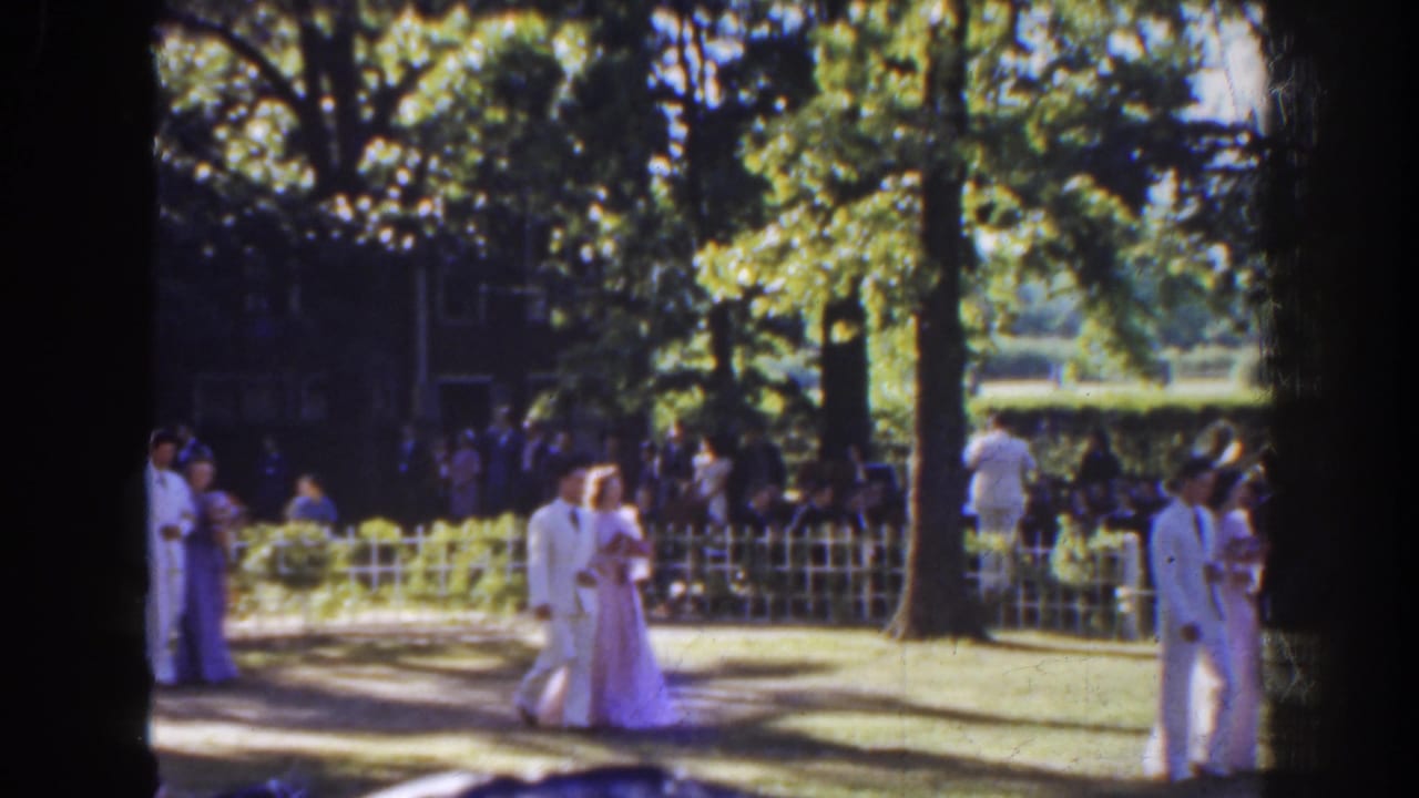 Couples in formal clothing walk across a shaded lawn while guests watch behind a white fence.