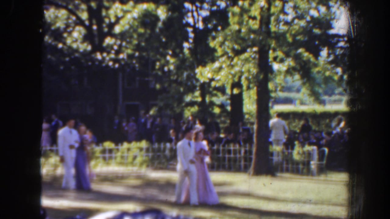 A couple walks together near the center of a lawn as guests stand behind a low white fence.