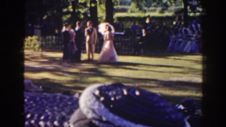 In 1941 Florida home-movie footage, a bride, groom, and young attendants gather on a sunlit park lawn with guests seated behind them.