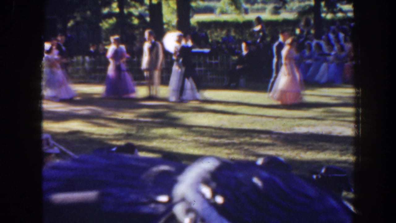 Bride and attendants stand near guests, with a parasol visible beside the group.