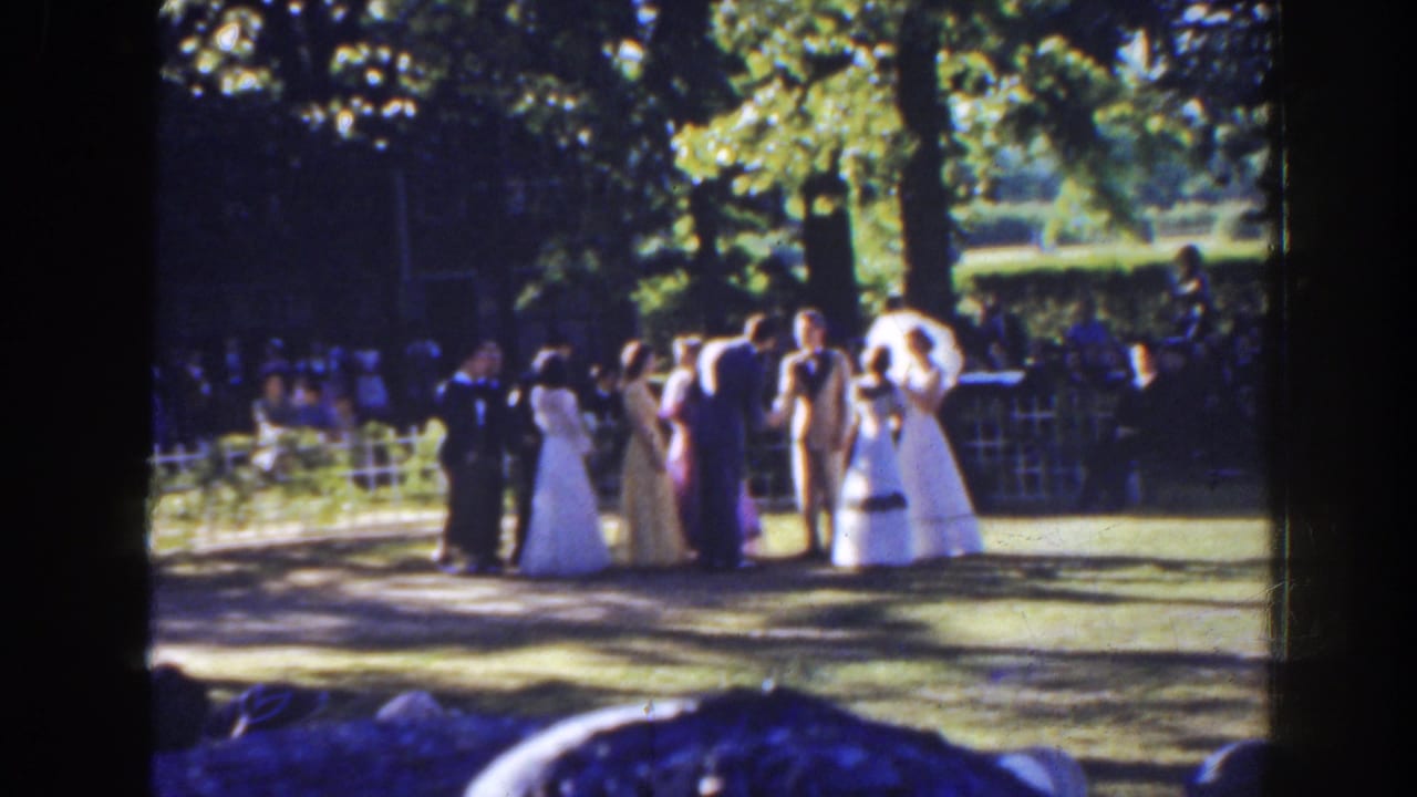 1941 Florida home-movie frame showing formally dressed guests gathered on grass under leafy trees, with onlookers behind a low fence.