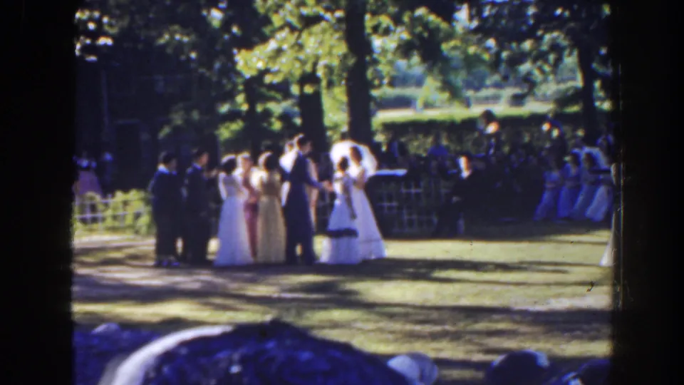 1941 Florida home-movie still of formal guests mingling on grass, with a parasol, shaded trees, and seated spectators in the background.
