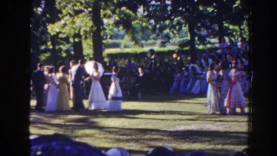 1941 Florida frame showing formally dressed guests spread across a sunny lawn, with women in light dresses, men in suits, parasols, and seated onlookers.