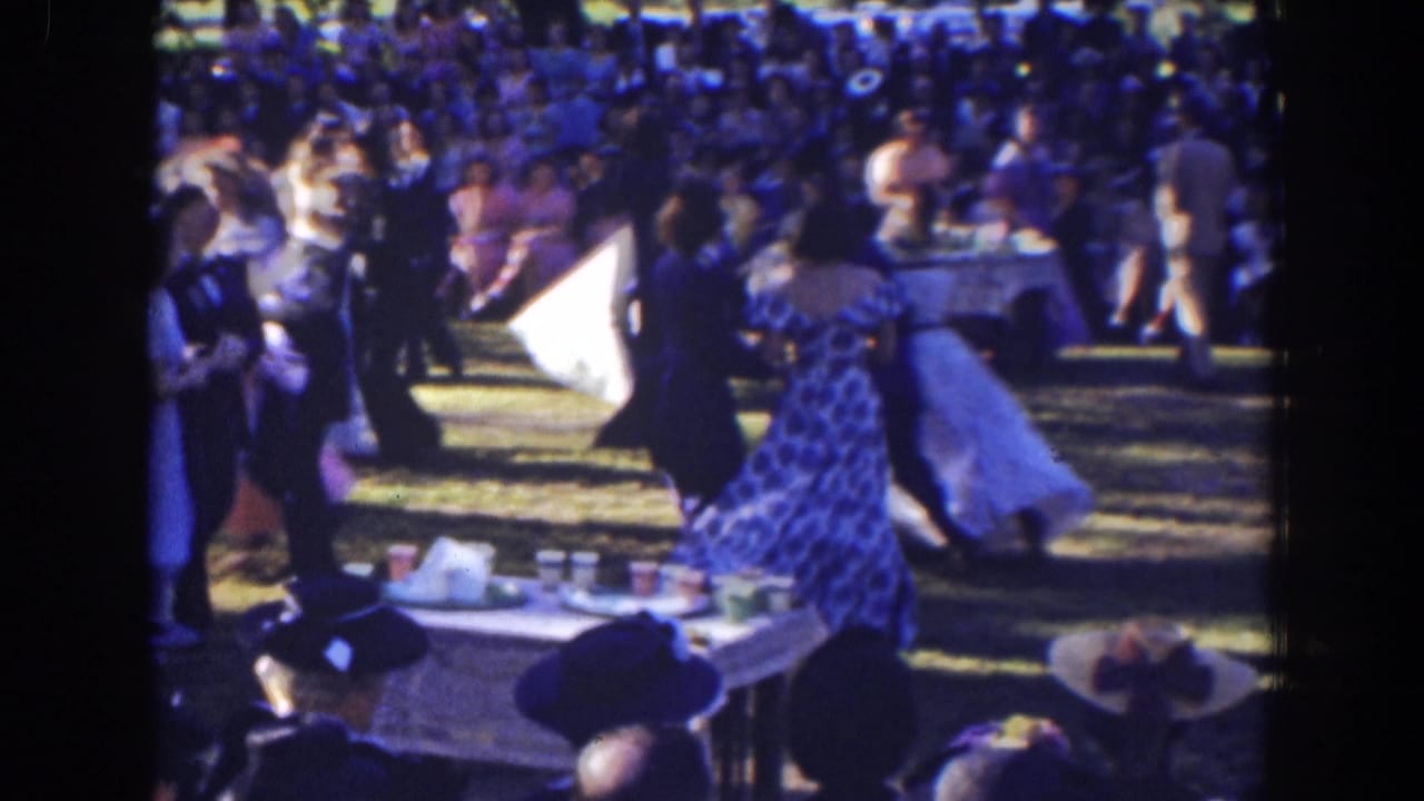 Dancers in long dresses and suits move past a table set with cups outdoors.