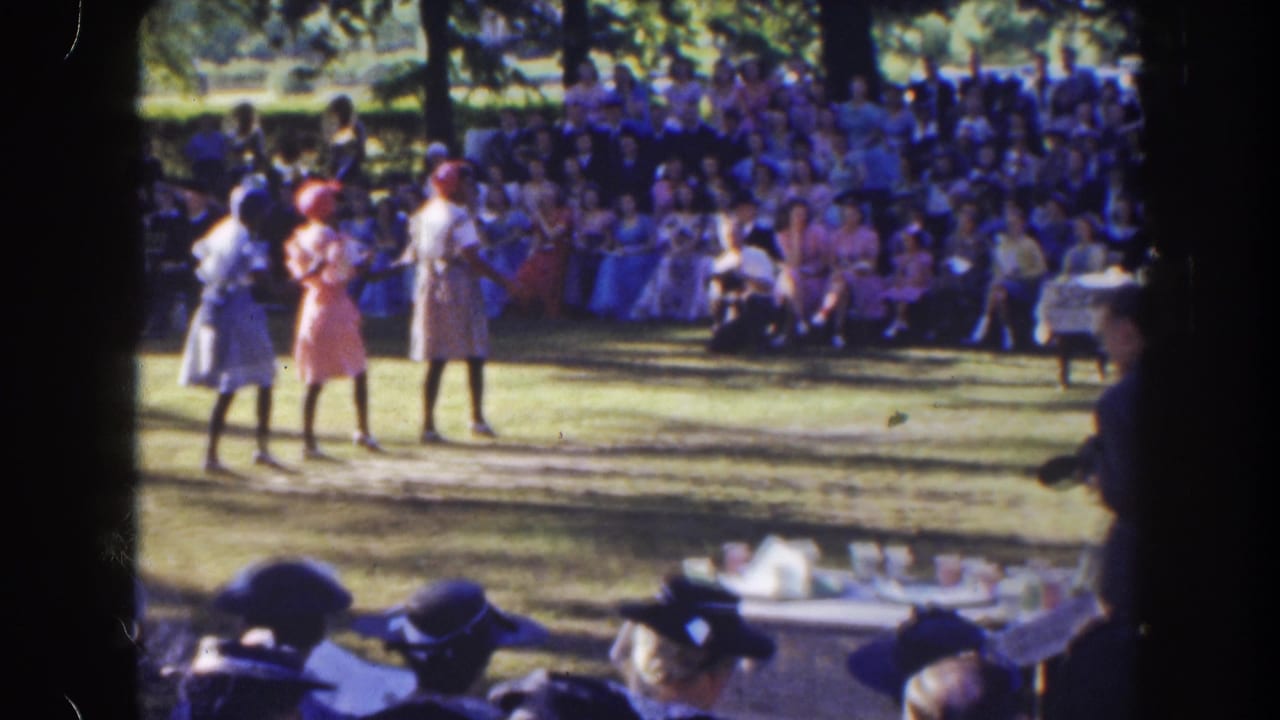 Three women stand on a sunny lawn before a seated audience.