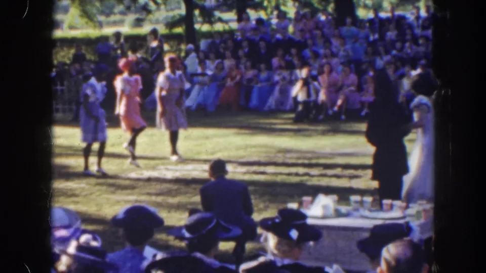The home-movie frame shows three women dancing on a lawn, watched by a large audience in 1940s-style clothing and hats.