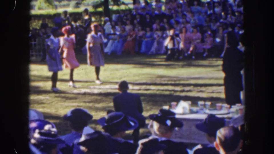 Three women stand in formation on a Florida lawn while a seated crowd watches, with hats and a table visible in the foreground.