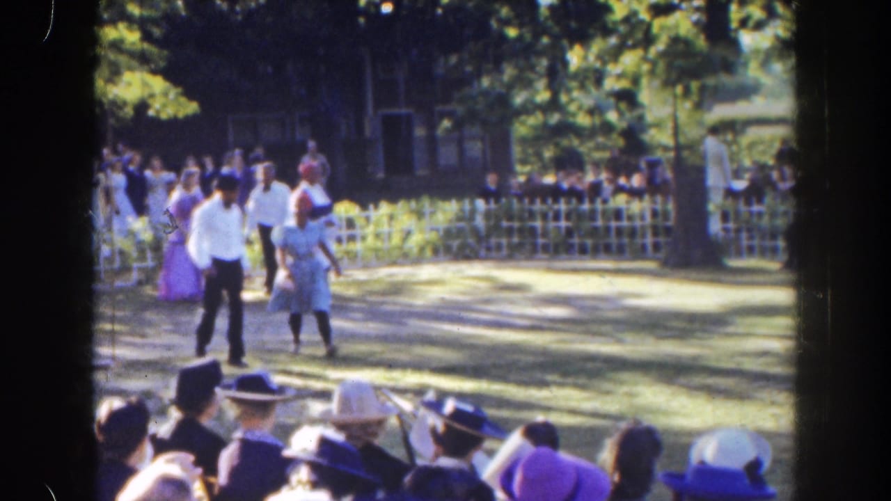 Four period-dressed people walk across a lawn while a seated crowd watches.