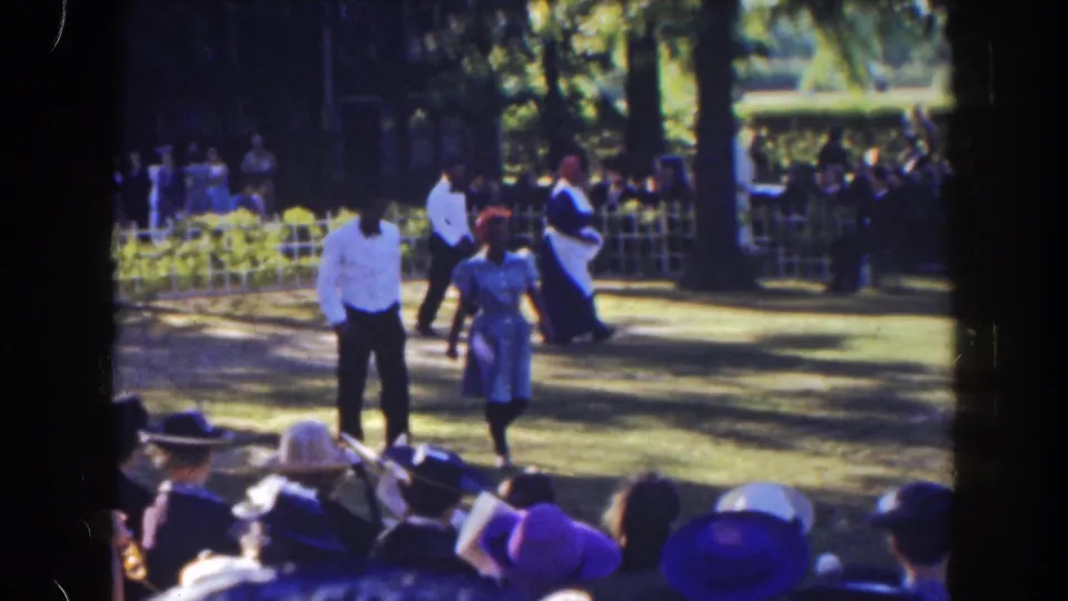 Two period-dressed walkers move closer to the camera while spectators sit in hats at the edge of the lawn.