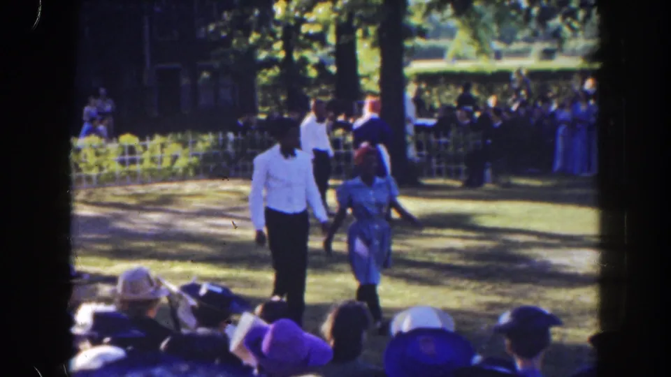 The group continues past the crowd in a sun-dappled Florida park setting, with hats and formal dress visible.