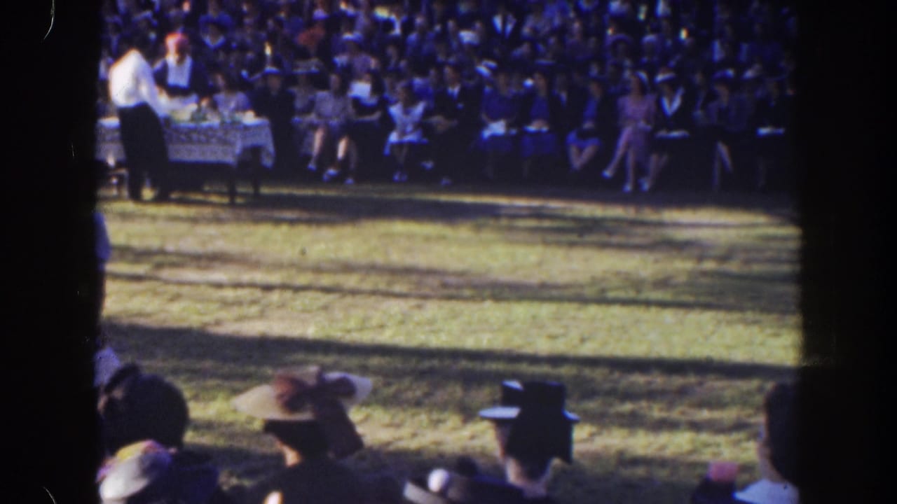A seated crowd watches a table set on a lawn under trees.