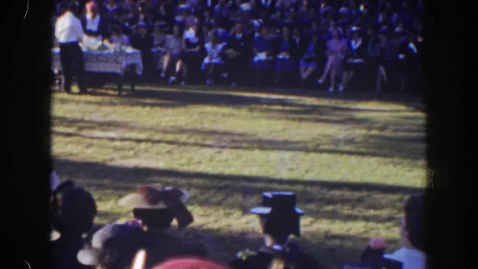 Foreground spectators in hats frame the view toward a formal table and seated audience at the 1941 Florida outdoor gathering.