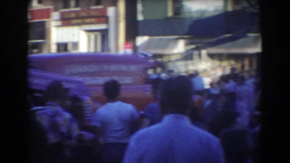 Crowd in 1958 York, Nebraska seen from behind as people look toward a red van and storefronts on a busy downtown block.