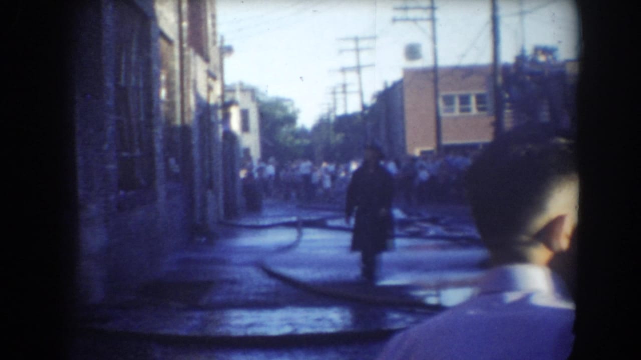 Man in dark coat on wet street with hoses, crowd and foreground bystander at right.