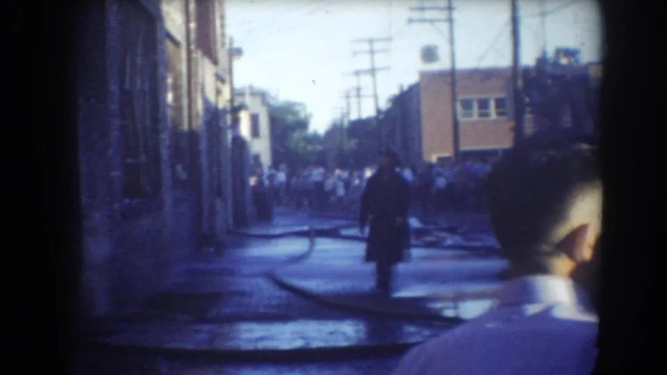 York, Nebraska street scene with a dark-coated man, fire hoses, bystanders and a red vehicle partly visible near the crowd.