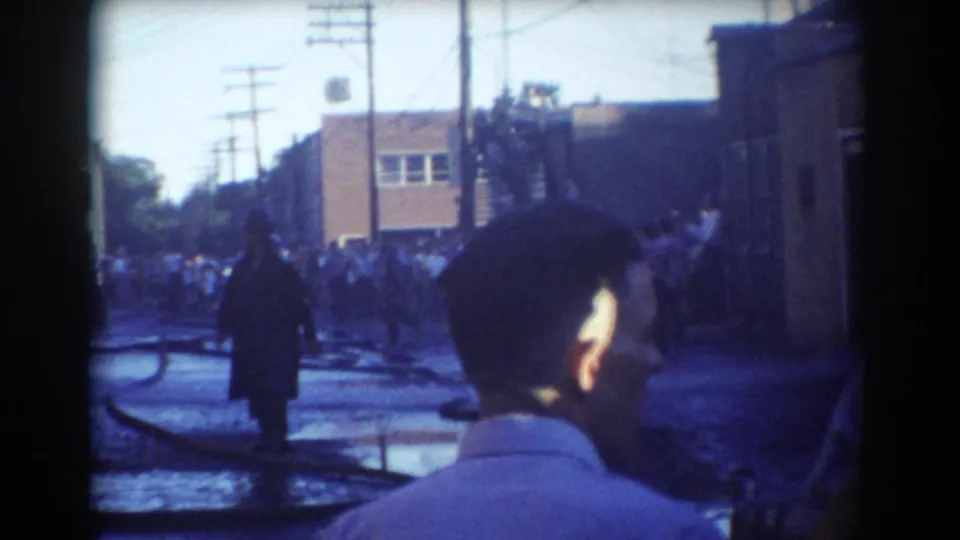 Home-movie view of a York, Nebraska fire-response street with a foreground spectator, wet pavement, hoses and a crowd in the background.